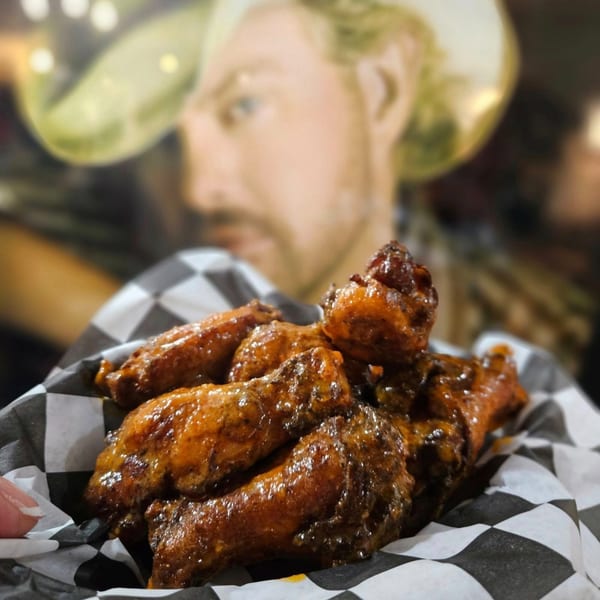 A basket of glistening, saucy chicken wings on checkered paper, with a blurred cowboy figure in the background.