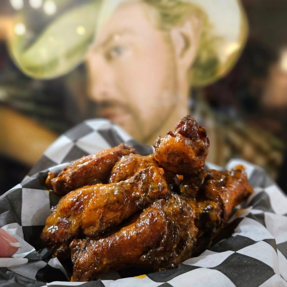 A basket of glistening, saucy chicken wings on checkered paper, with a blurred cowboy figure in the background.